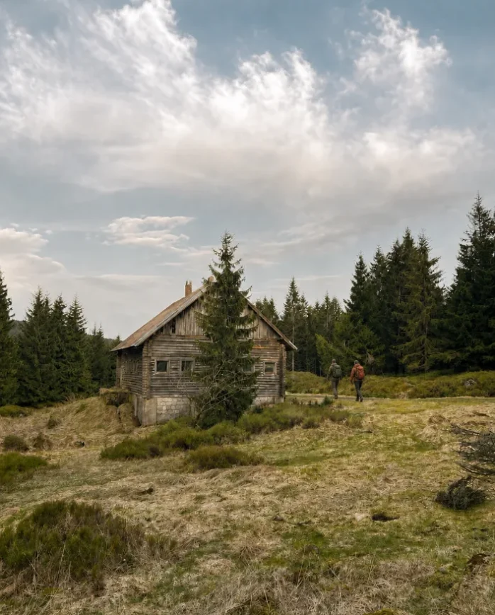 Dva turisté s batohy vyrážejí po horské cestě kolem dřevěné chaty v Jizerských horách.